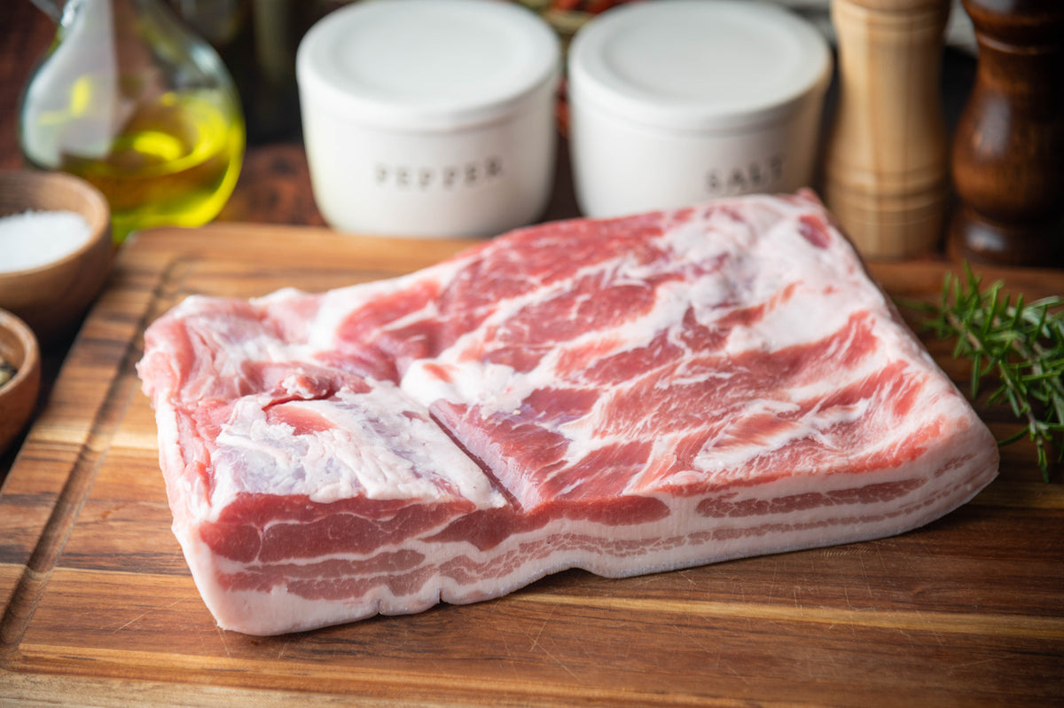A thick cut of raw marbled pork belly resting on a wooden cutting board, with salt and pepper shakers, a bottle of oil, and herbs in the softly blurred background.