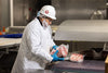 Person in a white lab coat and hard hat packaging meat in a industrial setting
